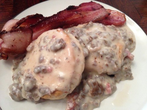 A plate of biscuits topped with creamy sausage gravy, accompanied by crispy bacon strips.