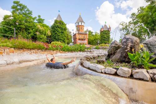 A person slides down a water ride on an inner tube, surrounded by lush greenery and a castle-like structure in the background.