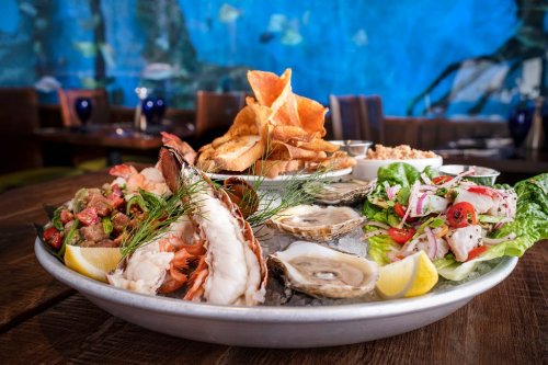 A seafood platter featuring shrimp, oysters, crab, and chips, garnished with lemon and fresh herbs, set against a blue backdrop.