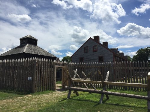 Historic fort with wooden palisade, two buildings, and a blue sky with clouds.