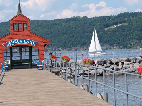 A red gazebo at Seneca Lake with a wooden pier, flowers, and a sailboat in the distance against a green hillside.
