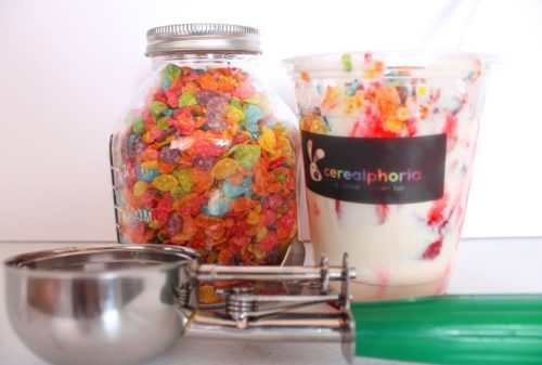 A jar of colorful cereal next to a cup of yogurt topped with fruit and cereal, with an ice cream scoop in front.