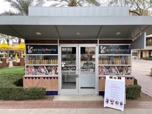 A small cereal bar kiosk with a glass front, featuring colorful cereal boxes and drinks, surrounded by palm trees.