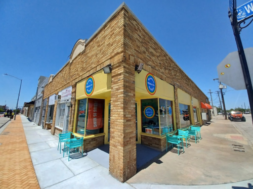 A corner storefront with yellow walls and large windows, featuring outdoor seating in a sunny urban setting.