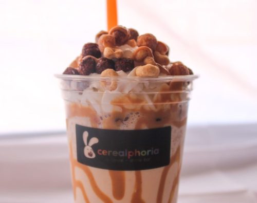 A cup of creamy dessert topped with cereal, nuts, and a drizzle of caramel, featuring a bright orange straw.