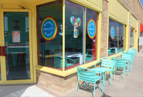 Brightly colored exterior of a dessert shop with rainbow-themed signage and turquoise outdoor seating.