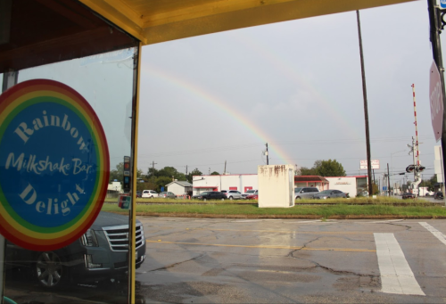 A view from inside a milkshake bar showing a double rainbow over a street with cars and buildings in the background.