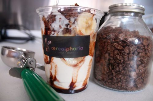 A cup of layered dessert with chocolate, bananas, and ice cream next to a jar of chocolate cereal and a scoop.