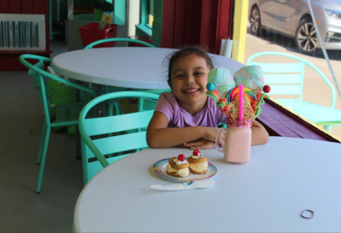 A smiling girl sits at a table with a colorful milkshake and cupcakes, enjoying a treat in a bright café.