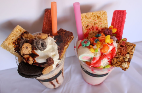 Two colorful milkshakes topped with whipped cream, cookies, cereal, and wafer snacks, served with pink straws.
