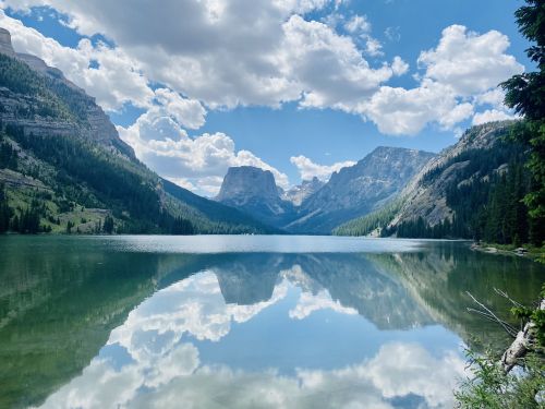 A serene lake surrounded by mountains, reflecting clouds and trees under a bright blue sky.