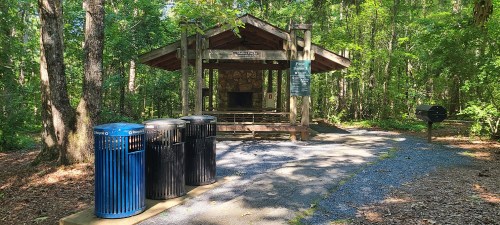 A picnic shelter in a wooded area with trash bins and a grill nearby, surrounded by trees and gravel paths.