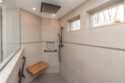 Modern shower with a rainfall showerhead, wooden bench, and large windows, featuring sleek tile walls.