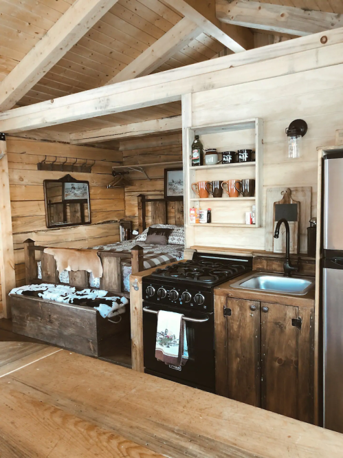 Cozy cabin kitchen with wooden walls, a stove, sink, and rustic decor, featuring a bed in the background.