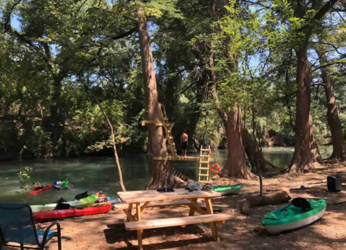 A serene riverside scene with kayaks, a picnic table, and trees, featuring a person climbing a tree in the background.