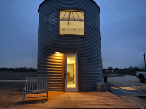 A rustic silo home at dusk, featuring a wooden deck, a rocking chair, and a large window above the entrance.