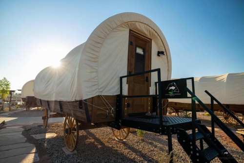 A vintage-style wagon with a rounded canvas top, set against a bright sky, featuring a wooden door and steps.