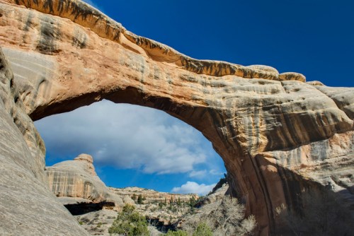 A natural stone arch rises against a blue sky, surrounded by rugged rock formations and sparse vegetation.