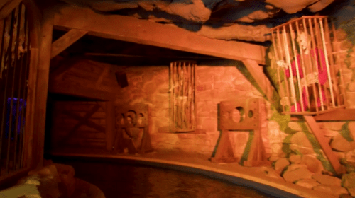 A dimly lit cave interior with wooden cages and a stone wall, featuring a water edge in the foreground.