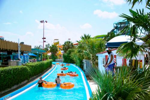 A lazy river at a water park with people in orange tubes, surrounded by palm trees and colorful slides in the background.