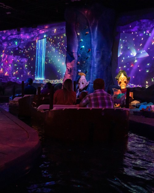 A dark, colorful indoor scene with a waterfall projection and whimsical figures, as people enjoy a boat ride.