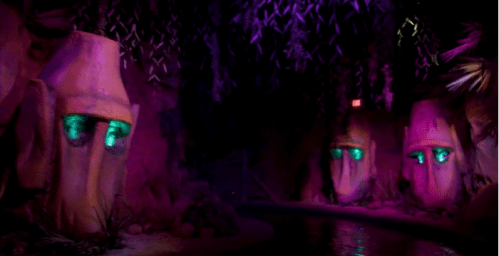 Colorful cave scene with two large, stylized faces and glowing green eyes, surrounded by lush foliage and water.