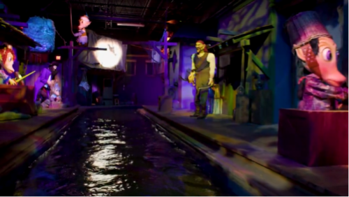 A colorful indoor scene with whimsical puppets and a waterway, illuminated by purple and blue lights.