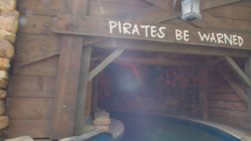 A wooden sign reading "Pirates Be Warned" above a dark waterway entrance, surrounded by rustic wooden walls.
