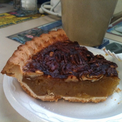 A slice of pecan pie on a white plate, with a brown mug in the background. The pie has a golden filling and a flaky crust.