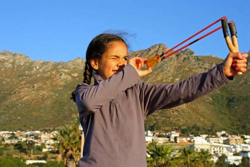 A girl aims a slingshot outdoors, with mountains and a town in the background, showcasing focus and determination.