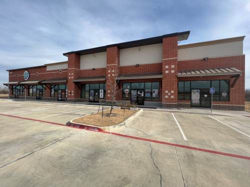 A brick commercial building with large windows and a parking lot, under a cloudy sky.
