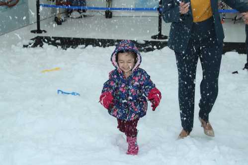 A joyful child in a colorful coat runs through artificial snow, smiling as snowflakes fall around her.