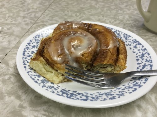 A cinnamon roll with icing on a decorative plate, accompanied by a fork, on a textured table surface.