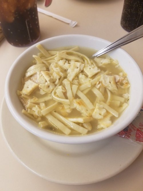 A bowl of chicken noodle soup with shredded chicken, noodles, and crackers, served on a plate with a spoon.