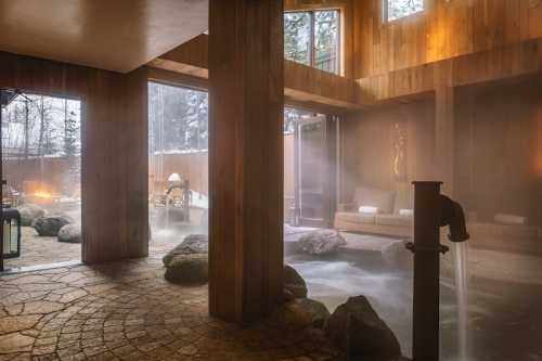 A serene indoor spa with wooden walls, large windows, and a misty atmosphere, featuring rocks and a water feature.