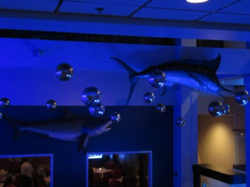 Two large fish sculptures hang from the ceiling, surrounded by reflective disco balls, in a dimly lit restaurant.