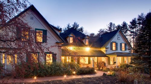 A charming two-story house with warm lights, surrounded by trees and a pathway, at dusk.