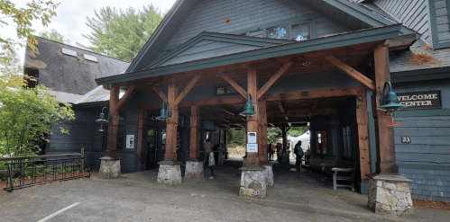 A rustic welcome center with wooden beams and stone accents, surrounded by greenery and people entering.