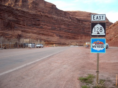 A road sign for East 128 and a Scenic Byway sign next to a dirt road surrounded by red rock formations.