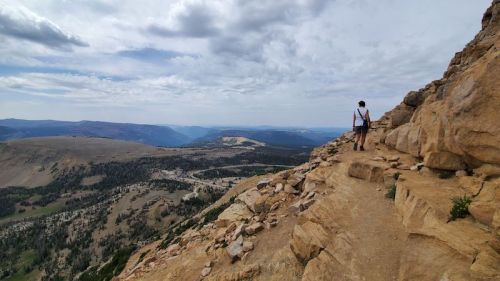 A person stands on a rocky mountain trail, overlooking a vast landscape of hills and valleys under a cloudy sky.