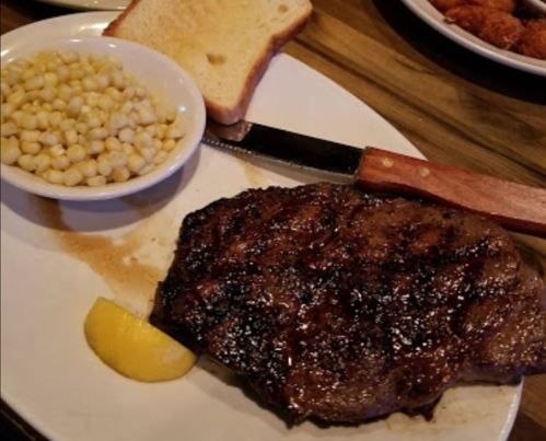 A plate with a grilled steak, a slice of bread, corn, and a lemon wedge, served with a knife on the side.