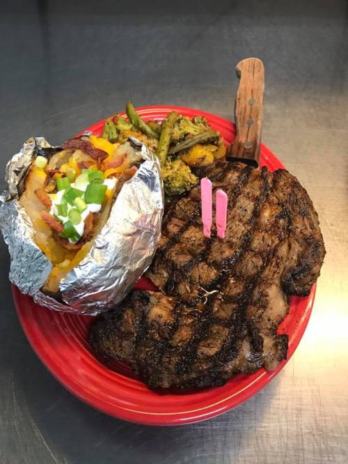 A grilled steak with a baked potato topped with bacon and green onions, alongside sautéed vegetables on a red plate.