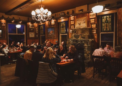 A cozy restaurant interior with wooden decor, diners at tables, and a stone fireplace, creating a warm atmosphere.