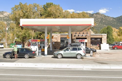 A gas station with two cars parked outside, surrounded by trees and mountains in the background.