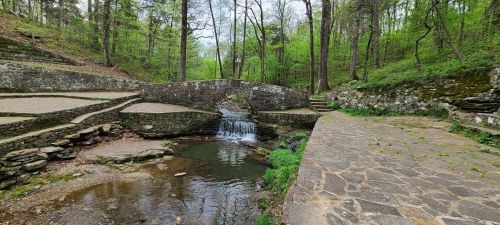 A serene forest scene featuring a small waterfall and stone pathways surrounded by lush green trees.