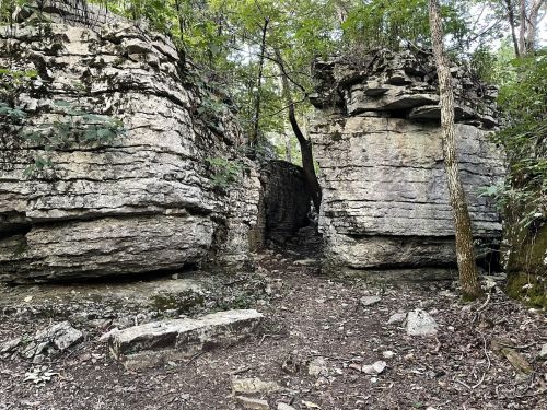 A narrow path between two large rock formations, surrounded by trees and greenery.