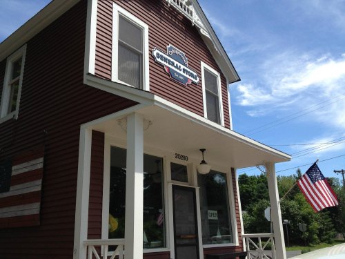 A red wooden building with a sign reading "General Store," featuring an American flag and a clear blue sky.