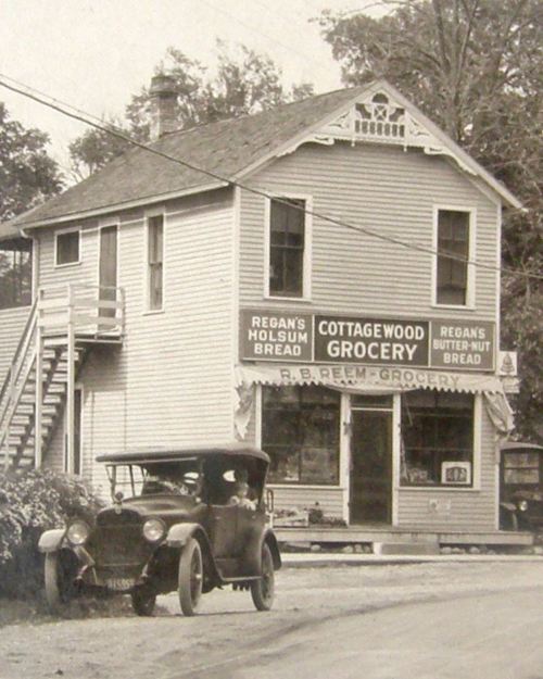 A vintage grocery store with a sign for "Cottagewood Grocery" and an old car parked in front. Black and white photo.