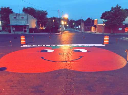 A large, smiling red face painted on a street, with traffic cones marking the area, under evening lights.