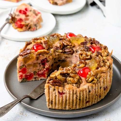 A slice of fruitcake topped with nuts and cherries on a gray plate, with more cake in the background.